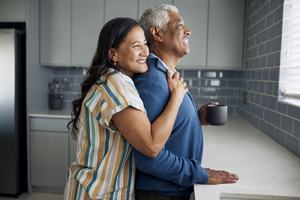 Elderly couple standing in a kitchen, smiling and hugging.