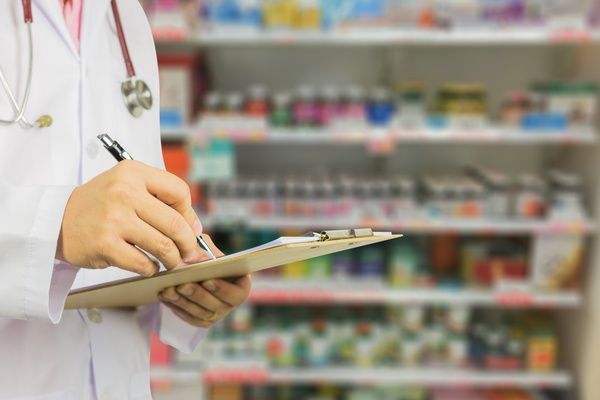 Blurred image of a doctor writing on a clipboard with shelves of medication in the background